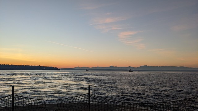 The sun sets behind the Olympics, with faint pink clouds in the sky, reflecting off the waters of Puget Sound. The car deck of the MV Kaleetan can be seen in the foreground.