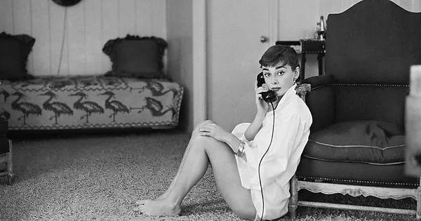Audrey Hepburn sitting on the thick-carpeted ground. She is only wearing a men's dress shirt (and some bracelets). This shows off her legs in the best possible way.  She's talking on the phone and looking toward the photographer with raised eyebrows. 