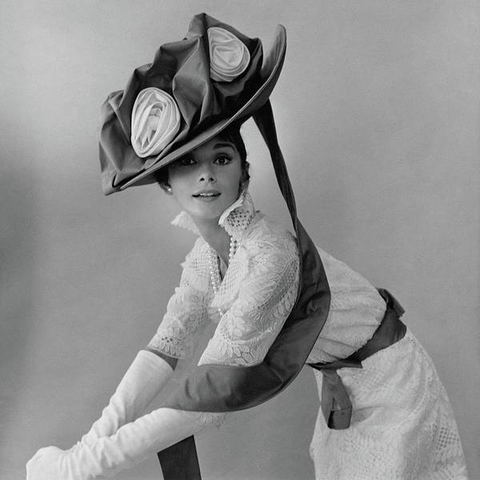 Audrey Hepburn in a costume from My Fair Lady - although I don't think it's from the movie. Anyway, there's a massive big hat with fabric bows and ribbons. She's leaning over and looking back at the camera. She's half-smiling with her mouth slightly open, looking slightly surprised. 