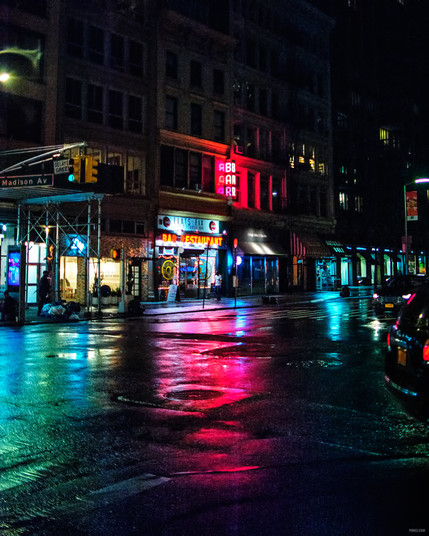 A cold, rainy night on the streets of New York City. The light from a bar reflects on the wet streets.