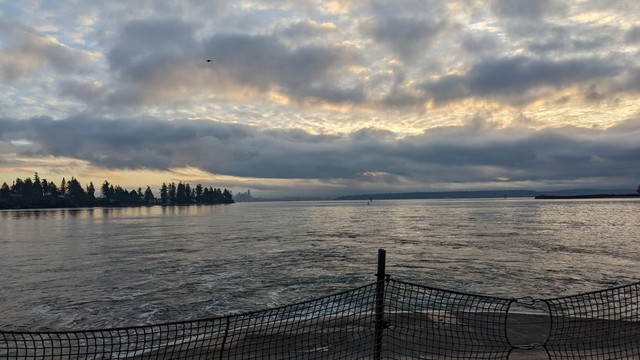 Light from the sun is mostly blocked by a layer of low clouds, which glow light orange. The sky reflects off the waters of Eagle Harbor. The net that closes off the number 1 end of MV Kaleetan's car deck is visible in the foreground.