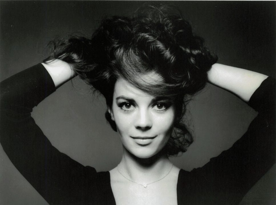 Portrait of Natalie Wood. She is looking right into the camera with a puckish grin. Her hands are above her head in her hair, pulling up up slightly into a lovely muss. 