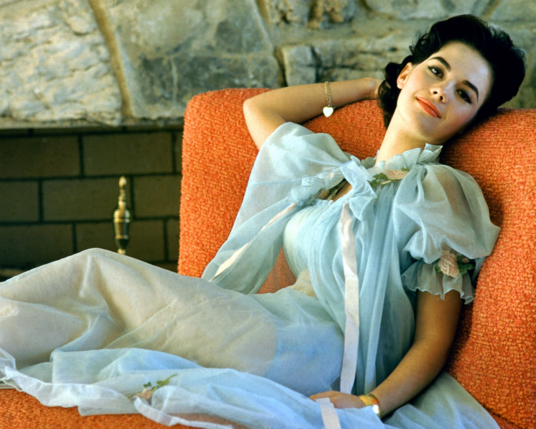 Natalie Wood reclines on a livid orange couch. Her head is tilted, laying on the back of the couch. She looks happily content. She is wearing an elaborate transparent pale blue negligee. 