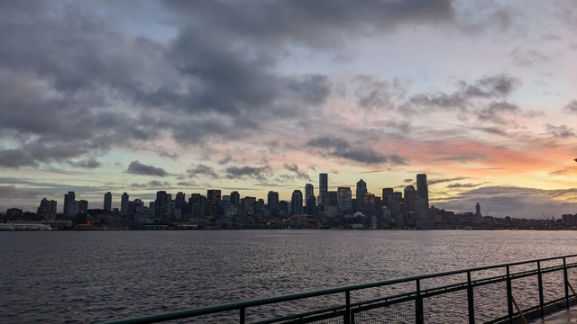 The sun rises behind clouds and the silhouetted city of Seattle, reflecting off the waters of Puget Sound as seen from a Washington State Ferry.