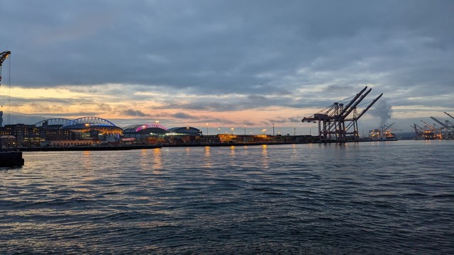 The sun rises behind the stadiums of Seattle, reflecting off the waters of Puget Sound. Cranes from the cargo terminal are in the distance on the right.