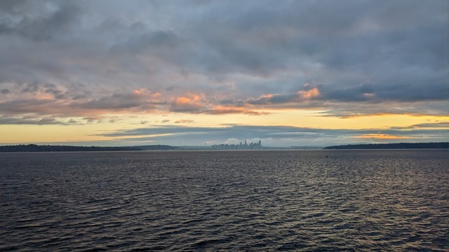Sunrise as seen from a Washington State Ferry coming out of Eagle Harbor on the way to Seattle. The sun is behind clouds, but shining orange through a gap, painting the bottom of some of the clouds red. Seattle is centered and small in the distance.