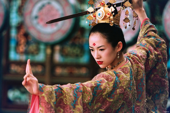 Zhang Ziyi in a still from House of the Flying Daggers. She is in a spectacular multi-colored dress and wears a golden crown with dangling decorations. She wields a sword above her head and gestures with her other hand.