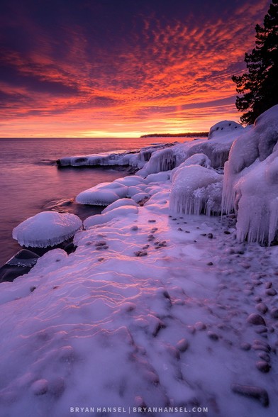 A vertical photo of an icy shoreline painted purple and orange by the setting distance sun on the horizon.
