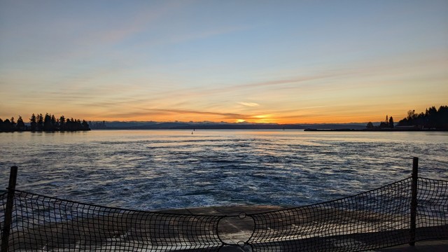 The sun rises in the distance, behind mountains shrouded in low clouds. The sky is mostly clear and blue, with a few whisps of faintly pink clouds. Silhouetted trees of Bainbridge Island are to the left and right of the frame, with the waters of Eagle Harbor in between. The car deck and safety net of a Washington State Ferry is in the foreground.