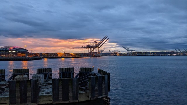 A patch of a cloudy sky glows pink-orange around the cranes of the Seattle cargo terminal, which reflects on the waters of Puget Sound. A "dolphin" structure that is part of the dock for a ferry is in the foreground.