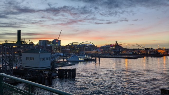 The sun rises behind the stadiums and shipping terminal of Seattle, Washington, reflecting on the waters of Puget Sound. The overhead walk of Slip 3 at Coliman Dock can be seen, and the green railing of the upper deck of a ferry is in the foreground.