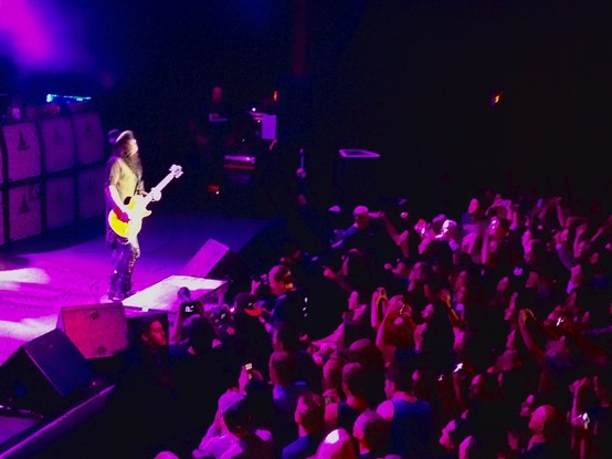 Slash, playing guitar on a magenta-bathed stage. He’s looking up as he plays, his leather top hat tipped slightly back. 