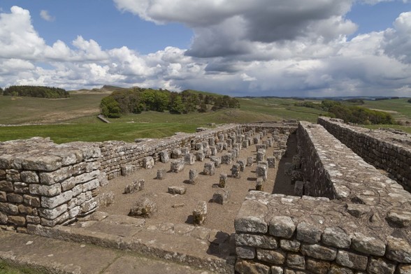 A bright day with plenty of white and grey clouds but large patches of clear blue sky too, over a typically English countryside view of rolling, grassy hills and groupings of trees. In the foreground are the ruins of a Roman building forming the outline of a long barn-like building with stone walls, complete and less than ten bricks high on three sides, but with an opening in front of our view. Dropping down from this entrance the ground space is filled with stone blocks arranged equidistant in a grid six wide by at least fifteen long.