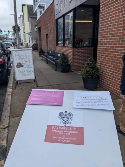 A donut box held in front of the viewer, with an out of focus store front behind. The box is labeled for DJ's Market & Deli and prominently features the eagle of the Polish coat of arms. The store front has a sandwich board out front reading "Paczki Day! Order yours today!"