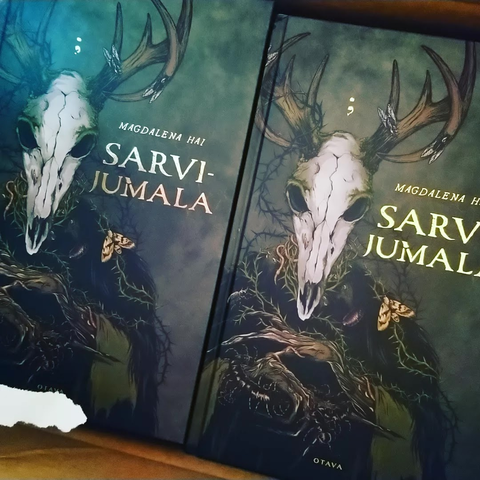 Two copies of Sarvijumala novel side by side in a box. The cover is greyish green with golden title and a deer skulled monster holding insects in his skeletal hands.