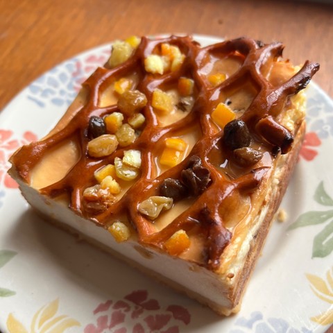 A big square of baked cheesecake on a floral plate. It has a thin pastry base, a pastry lattice top, lemon glaze and many marinated sultanas and citrus candied peel dotted through and scattered over. 