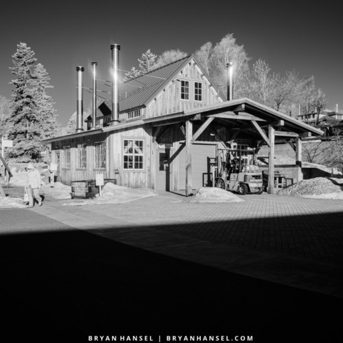 A black and white infrared image of a big shadow in teh foreground and a wooden blacksmith shop in the background.