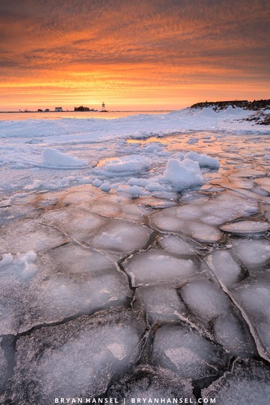 A vertical photo of sunrise over cracked ice that leads to a large body of water and a lighthouse. The orange and yellow sunrise is reflecting in the ice.
