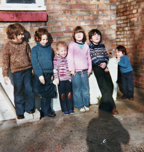 Close to the back of a brick house with its lower seven rows of bricks painted white. To the right of the house is an alleyway leading to the front of the house out of view. Standing against the wall are five children, all under ten, and there is another boy in the alleyway, just looking in. From left to right there are two boys - clearly twins, distinguishable only by their jumpers which are brown and blue respectively, then two girls - one younger than the other, then a boy with arms folded in front of him, then the youngest boy in the alley. Everyone has jeans and jumpers on and everyone is squinting as they're looking into the sun. The shadow of the photographer stretches across the concrete floor towards them all.