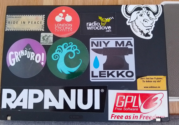 Photograph. One black laptop, its lid seen from the top. A series of stickers on the laptop lid - some newer, some older.
