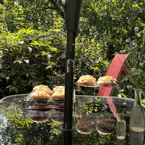 Pic taken from my reading chair. In the middle is the sturdy pole of the big parasol. Behind that the cooling racks wiyj the onion breads. Behind those the still lusciously green hedge. In front of the parasol pole are two small vases with wild flowers. The one on the left holds some bluish-purple flowers; the flowers in the right vase are a dirty white, but there's also a twig with red berries in that one. 