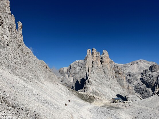 Looking back at Rifugio Re Alberto 1, with the Vajolet towers behind it.
The rifugio is sided by ragged mountain peaks; a hiking path crosses the scree slope at the bottom of one side of the peaks.