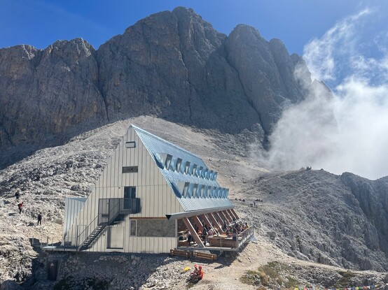 Rifugio Santner, at 2734m altitude. A metal triangular prism lying on its side with a terrace overlooking the chasm in front of it. A cloud rises up the cliff side.