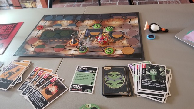 Sinbad (orange base), Medusa (green base), and three Harpies have surrounded Willow (played by a red tea pot) on the Unmatched game board. Angel (played by a penguin) is lying next to the board in defeat. In the foreground you can see cards from both Sinbad and Medusa. The lucky Gaze of Stone card, which dealt a whopping 10 damage to Willow, is on the top of Medusa's discards.