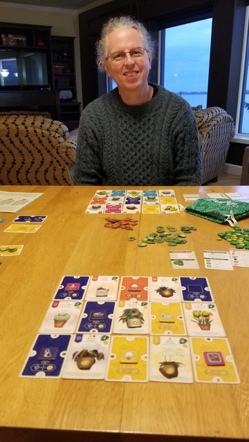 Peter smiles at me from across the gaming table. His 5 X 3 grid of diverse room and plant cards is in front of him. My grid is in the foreground. Most of my rooms were blue and yellow with a satisfying number of matching items. A scattering of pots, green thumbs, and leaves are in the middle of the table between us.