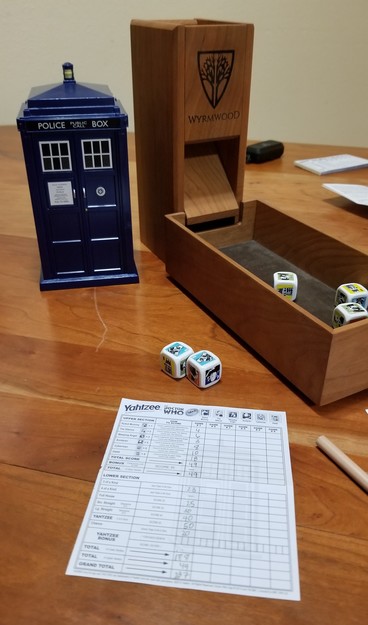 A fancy wooden dice tower sits on a table. Three dice are in the tray with two more on the table. The dice have pictures of six different Doctor Who villains on their sides. The TARDIS-shaped game box is sitting next to the tower with the completed score sheet in the foreground.