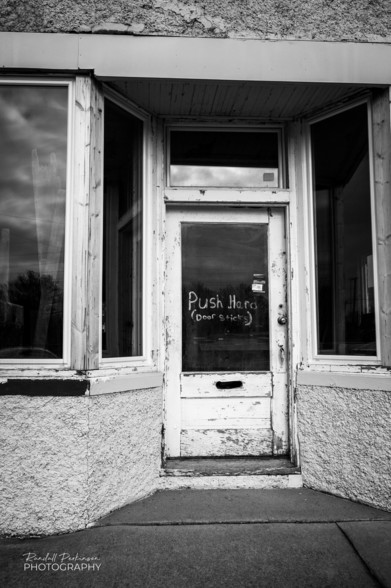 Old weathered door into retail space with windows on either side. Handwritten note on door reads, "Push Hard. Door Sticks." 