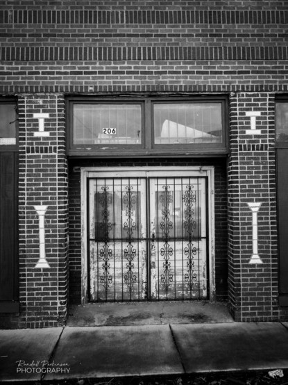 Brick building with a pair of white doors with ornate black iron gates in front of them. Either side of door has white glazed brick paaterns inset into brick columns. 