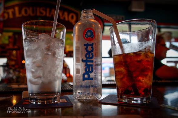 A glass of water, a glass of diet Dr. Pepper, and a plastic water bottle with the word "Perfect" on it sit on a bar. 