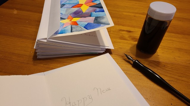 A stack of cards, a jar of ink, and a metal nibbed dip pen are laid out on a wooden surface.