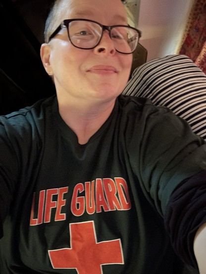 An enby person with very short hair and big glasses and not quite smiling for a selfie, wearing a dark green tee shirt with an orange lifeguard cross and lettering that says life guard. Lettering out of the frame reads: northshore.hi