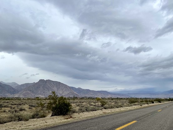 Wide landscape photo of a highway running through the desert with yellow-striped asphalt in the foreground; sand & scrub & desert plant in the mid-ground; & purple, navy, grey mountains against a huge dark sky with heavy rain clouds