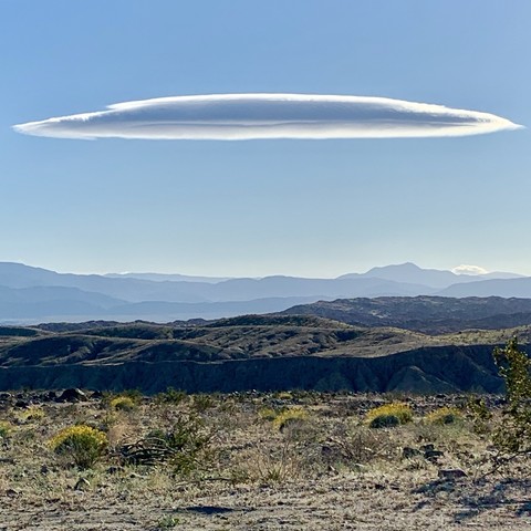 Landscape photo of a lenticular cloud (that looks like a flying saucer) in a big, blue sky over mountains & desert sand, rock, grass, & scrub