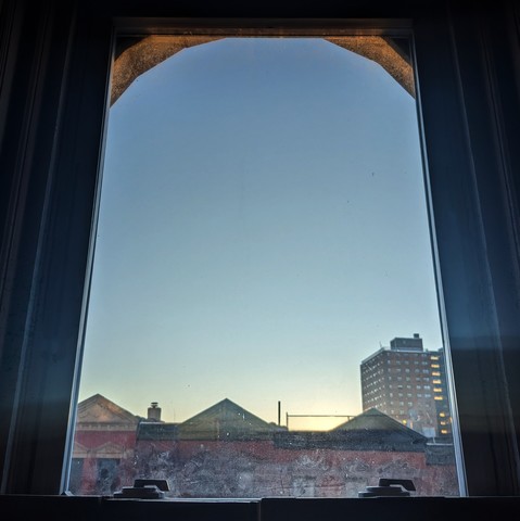 Looking through an arched window twenty minutes after sunrise the cloudless sky fades from parchment at the horizon to celeste above. Pointed roofs of Harlem brownstones with red brickwork are across the street, and a taller apartment building can be seen in the distance. The window is dirty and showing grime on the bottom. The top arch of the window is lit up orange from the angle of the sun.