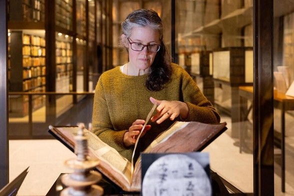 A woman carefully turns a page in a large bible inside a large glass display case. In the foreground are objects with Chinese characters. In the far background are rows of book shelves and more display cases.