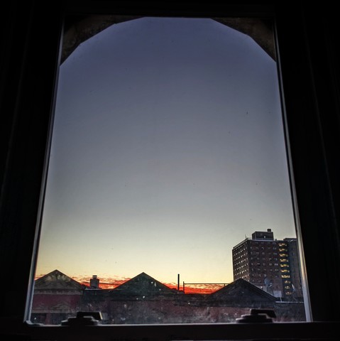 Looking through an arched window twelve minutes before sunrise the sky fades from pale yellow at the horizon to pale blue above, with thin streaks of deep orange at the skyline. Pointed roofs of Harlem brownstones with red brickwork are silhouetted across the street, and a taller apartment building can be seen in the distance. 