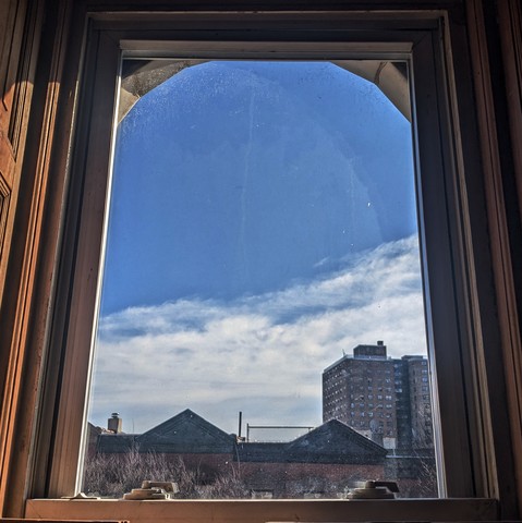 Looking through an arched window during late morning, the bottom half of the sky is full of weak white clouds, and the top half is clear sky blue. Pointed roofs of Harlem brownstones with red brickwork are across the street, and a taller apartment building can be seen in the distance.