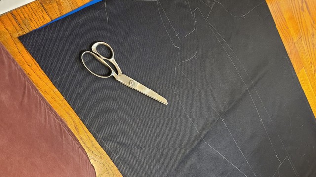 A large pair of scissors on a length of dark fabric with a coat pattern penciled onto it.