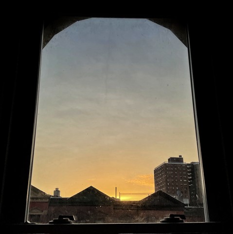 Looking through an arched window ten minutes after sunrise the sky is full of subtle watery clouds, fading from orange at the horizon to robin-egg blue above. Pointed roofs of Harlem brownstones are silhouetted across the street, and a taller apartment building can be seen in the distance. 