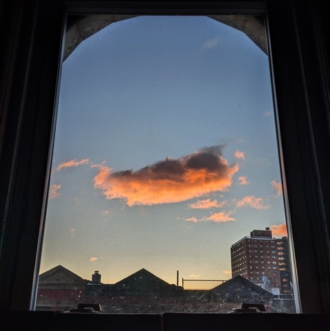 Looking through an arched window four minutes after sunrise there is a single large fluffy underlit plink cloud in the center of the blue sky. Pointed roofs of Harlem brownstones with red brickwork are across the street, and a taller apartment building can be seen in the distance. 