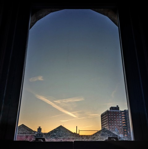 Looking through an arched window five minutes after sunrise the sky fades from orange to dark blue, criss-crossed by lines of clouds at the horizon. Pointed roofs of Harlem brownstones are silhouetted across the street, and a taller apartment building can be seen in the distance. 