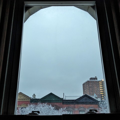 Looking through an arched window at the moment of sunrise the sky is a dense mass of clouds as a gentle snow falls. Pointed roofs of Harlem brownstones with red brickwork are across the street, and a taller apartment building can be seen in the distance. The tops of snow-covered trees are at the bottom of the frame. 