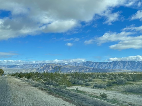 Wide shot of a beautiful blue & grey mountain range under a bright blue sky with clouds, taken from the roadside, surrounded by desert sand, shrubs, & grass