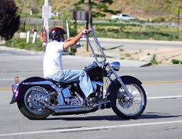 A side profile of a chopper motorcycle, mostly gleaming chrome with black tires and fenders. The rider is wearing a black helmet, a white T-shirt, light-colored blue jeans, and black shoes. Because it's a chopper, the handlebars are above eye level, so his arms extend forward from his shoulders at an upward angle to grasp them. 