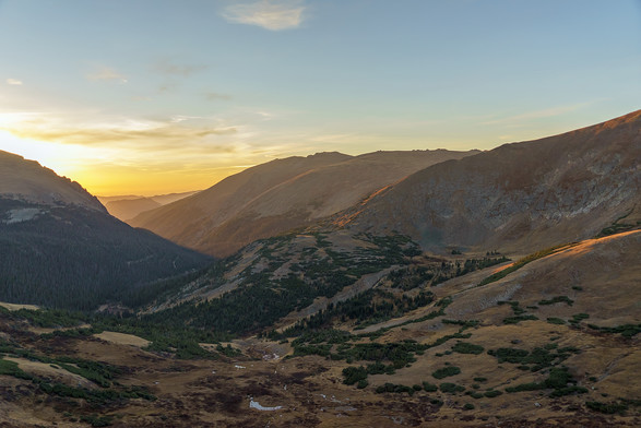Landscape photo. View from the Trail Ridge Visitors Center in Rocky Mountain National Park, looking east down into the Fall River Valley at sunrise. The valley below is becoming light enough to see details of the terrain, with small ponds and pools reflecting brightly. There is haze in the sky, blurring details of the hills at the horizon, glowing yellow in the light of the sun coming up behind the flank of mountain at the left of the frame. A ridge at the right and another in the center are sh…