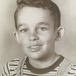 Closeup of my bio father's school photo showing his cute little 1950s boy face. His hair is combed just so, & he has lots of freckles, a turned up nose, & a lopsided smile. He's wearing overalls & a striped tee shirt.
